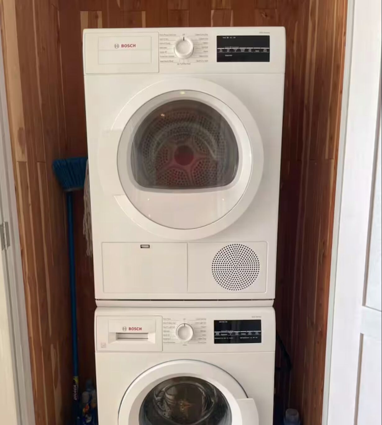 A stacked Bosch washing machine and dryer unit in a laundry room with wooden walls and a broom leaning against the wall.