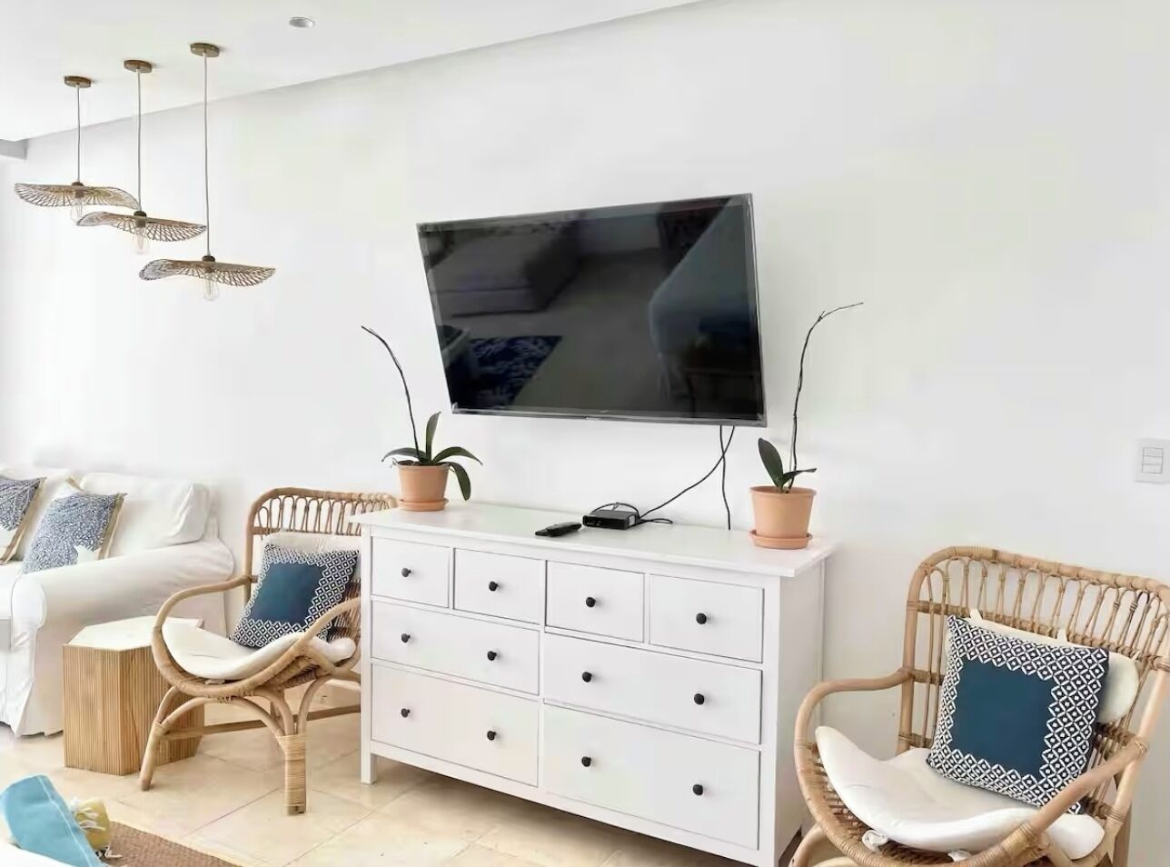 Een moderne woonkamer met een flatscreen televisie aan de muur, een witte kast met lades, twee rieten fauteuils met kussens en een kleine ronde tafel.