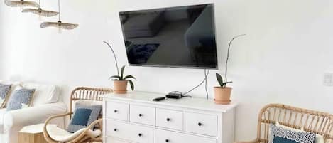 Een moderne woonkamer met een flatscreen televisie aan de muur, een witte kast met lades, twee rieten fauteuils met kussens en een kleine ronde tafel.