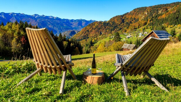 Deux fauteuils en bois, une bouteille de vin et des verres posés sur une souche d’arbre, le tout mis en valeur par un paysage de montagnes et d’un village en arrière-plan.