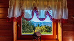 Une cabane en bois avec une fenêtre ornée d’un rideau en dentelle rouge et blanche, une petite maquette de maison en bois, et une vue sur une forêt et des montagnes.