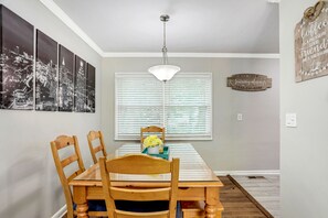 Un coin repas avec une table à plateau en verre, des chaises en bois, un luminaire suspendu, une œuvre d’art représentant un paysage urbain, une fenêtre avec des stores et une enseigne en bois accrochée au mur.