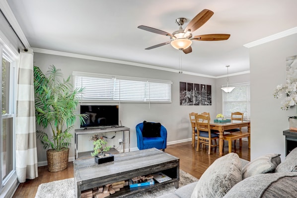 Ein Wohnzimmer mit Fernseher, blauem Sessel, Esstisch und Deckenventilator.