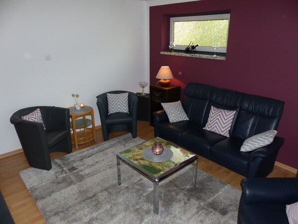 A living room with a dark blue sofa, two armchairs, a coffee table, a small side table with a lamp, and a window with a view of greenery. - Beautiful modern barrier-free bungalow in Rendsburg (Rendsburg)