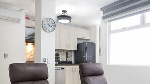 A modern living room with two brown leather chairs, a small table, and a grey rug. A kitchen is visible in the background with white cabinets and a black refrigerator.