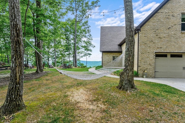 A hammock between trees in a grassy area with a view of a lake and a stone building with a garage.