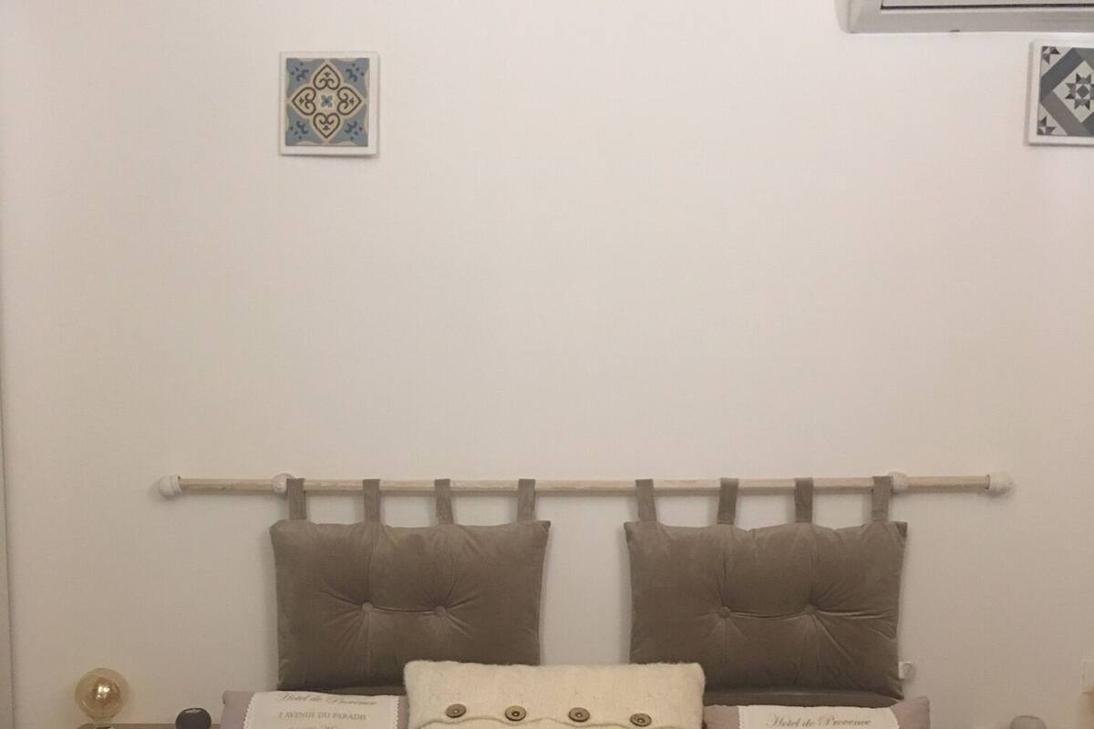 Una cama bien hecha con una manta que dice 'Home Sweet Home', dos almohadas con el texto 'Hotel du Provence', una mesita de noche con una lámpara y un aire acondicionado de pared.