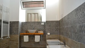 A bathroom with a large sink, a bidet, and a toilet. There is a towel rack with two towels and a bottle of lotion. The walls are tiled with a patterned design. - Dimora Castelli (Palermo)