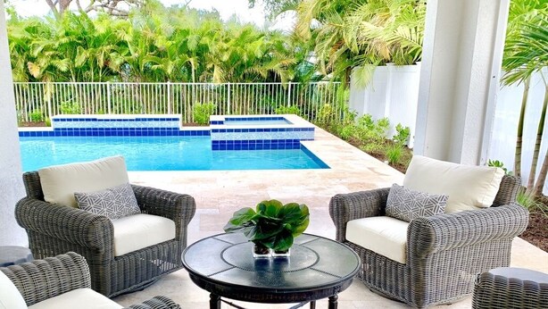 A poolside area with wicker furniture and a small table, surrounded by a white fence and lush greenery. - New Resort style Pool/Spa minutes to Vanderbilt beach! BlueBeachBungalow1 (Naples)
