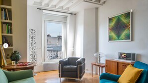 Un salón con un sillón de cuero negro, un sofá azul verdoso con un cojín amarillo, una mesita de madera, un aparador de vidrio y un ventanal con vistas a edificios.