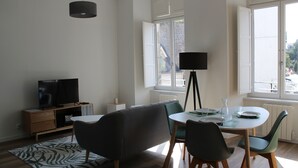 A modern living room with a dining area, featuring a sofa, a TV, a dining table with chairs, and a large window providing natural light.