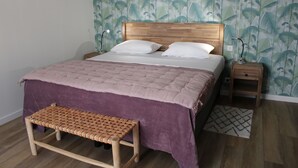 A bedroom with a wooden headboard, a purple bedspread, two white pillows, a woven bench, a nightstand with a lamp, and a patterned rug.