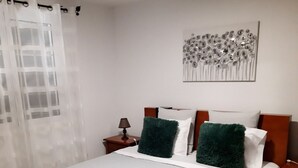 Ein ordentlich bezogenes Bett mit einer grün-weißen Tagesdecke, zwei grünen Kissen und einem weißen Kissen. Ein kleiner Nachttisch mit einer Lampe steht daneben, über dem Bett hängt ein Wandbild und ein Fenster mit durchsichtigen Vorhängen.