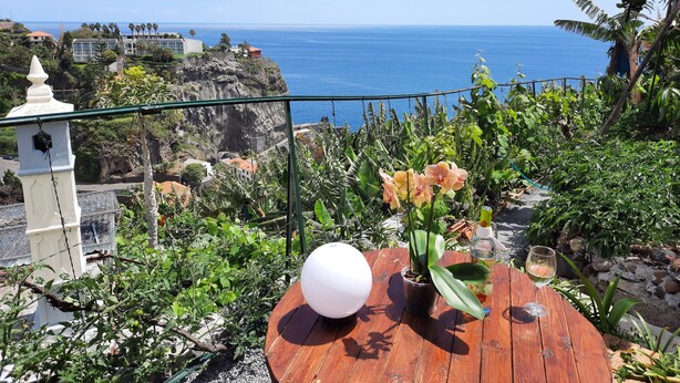 Ein Holztisch mit einem Blumentopf und einem Weinglas, mit Blick auf die Küste, ein weißes Gebäude und eine Klippe.