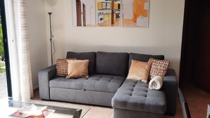 Ein modernes Wohnzimmer mit einer grauen Eckcouch, einem Glastisch und einem Flachbildfernseher. An der Wand hängen drei gerahmte Kunstwerke, eine Stehlampe und ein großes Fenster mit Vorhängen.