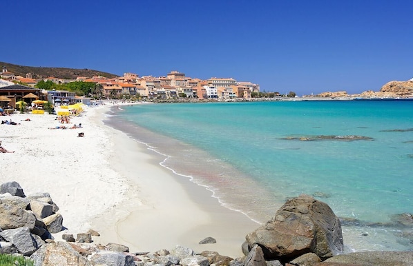 Una playa de arena blanca, agua turquesa cristalina y edificios al fondo.
