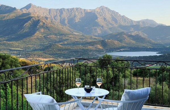 A balcony with a table and two chairs, overlooking a mountainous landscape with a lake.