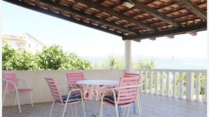 A covered patio with a table and chairs, a white railing, and a view of greenery and buildings.