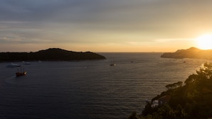 Escena costera con barcos sobre aguas tranquilas, un paisaje montañoso y una puesta de sol.