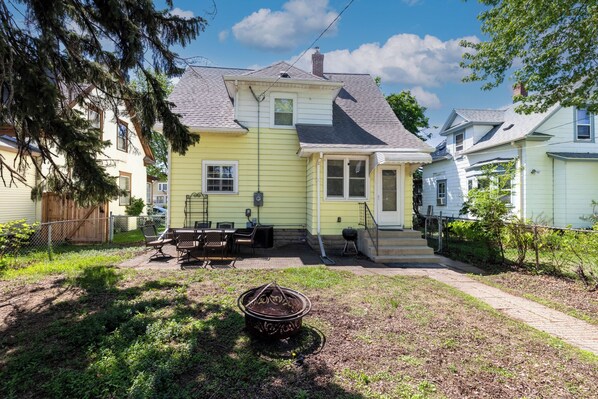 A two-story house with a front porch, a small backyard with a grill, and a fire pit. - 5BR Farmhouse Near Allianz Field • Theater • Backyard (St Paul)