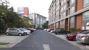 A parking lot with cars parked in rows, a building with multiple floors and windows, and a large billboard in the background. - D condo Hyde Chiangrai. Authentic Thai hospitality. Room service is provided. (phan)