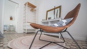 Een kamer met een rieten stoel, een kamerbreed tapijt met dessin, een wit schap en een spiegel aan de muur.
