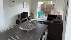 Ein modernes Wohnzimmer mit einem Flachbildfernseher, einer dunklen Couch, einem Esstisch mit dekorativer Schale und einer Glasschiebetür zu einem Außenbereich mit Holzzaun und Stühlen.