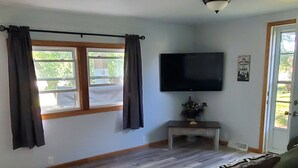 A living room with a flat-screen TV, wooden flooring, a sofa, and a window with curtains.