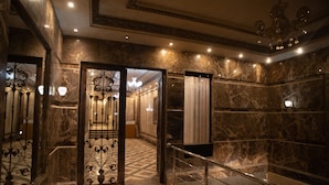 Un pasillo con piso de mármol, un candelabro, una puerta de vidrio con marco de metal decorativo y una escalera con barandilla de metal.