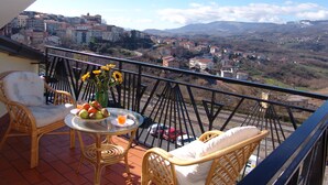 A balcony with wicker furniture, a glass table with fruit and a drink, and a view of a town and mountains. - B&B Il Tulipano - Rogliano (Calabria) Italy (Rogliano)