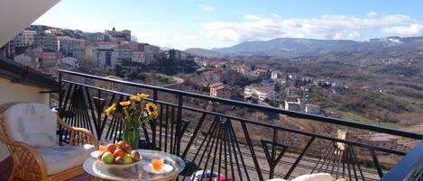A balcony with wicker furniture, a glass table with fruit and a drink, and a view of a town and mountains.