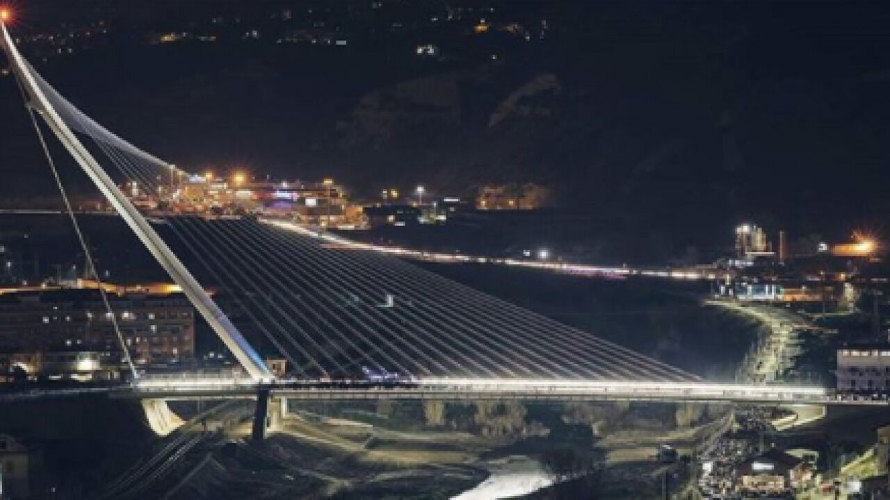 Een nachtzicht van een kabelbrug met verlichte kabels, over een waterlichaam met een stadsgezicht op de achtergrond.