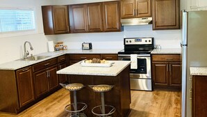 A modern kitchen with wooden cabinets, a stainless steel refrigerator, a built-in oven, and a bar area with stools. - Kuby's Place, Downtown Shiner Casita (Shiner)