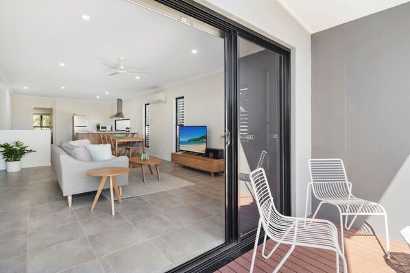 A modern living room with a sofa, coffee table, and TV. There is a sliding glass door leading to a balcony with two white chairs.