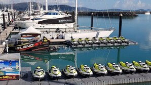 Un puerto deportivo con barcos amarrados y motos acuáticas alineadas en el muelle.