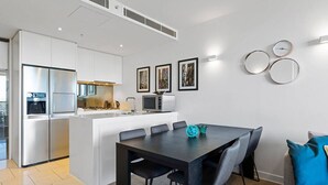 Une cuisine moderne avec des meubles blancs, un réfrigérateur en acier inoxydable et un coin repas comprenant une table et des chaises aux couleurs sombres.