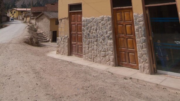 Um edifício rústico com uma placa que diz "Hostal". Apresenta paredes de pedra, portas de madeira e uma vista desobstruída de uma montanha ao fundo.