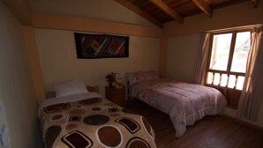 Quarto com duas camas, piso de madeira, uma janela grande com cortinas e uma tapeçaria colorida na parede.