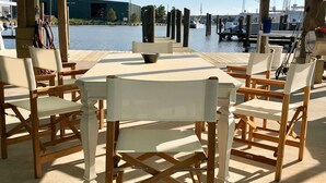 Un espace extérieur aménagé avec une table et des chaises, offrant une vue sur une marina avec des bateaux et un bâtiment en arrière-plan.