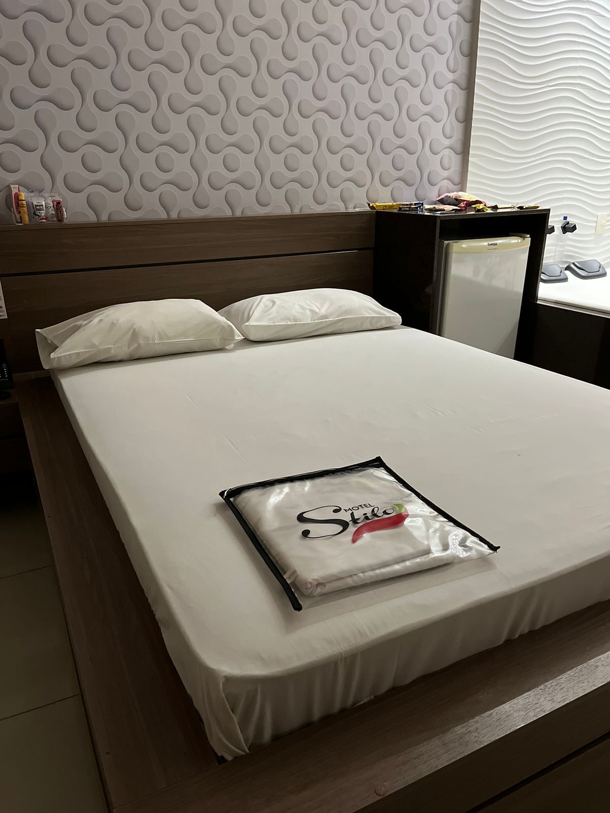 A neatly made bed with white sheets and pillows, a small bedside table with a lamp, and a refrigerator in the corner.
