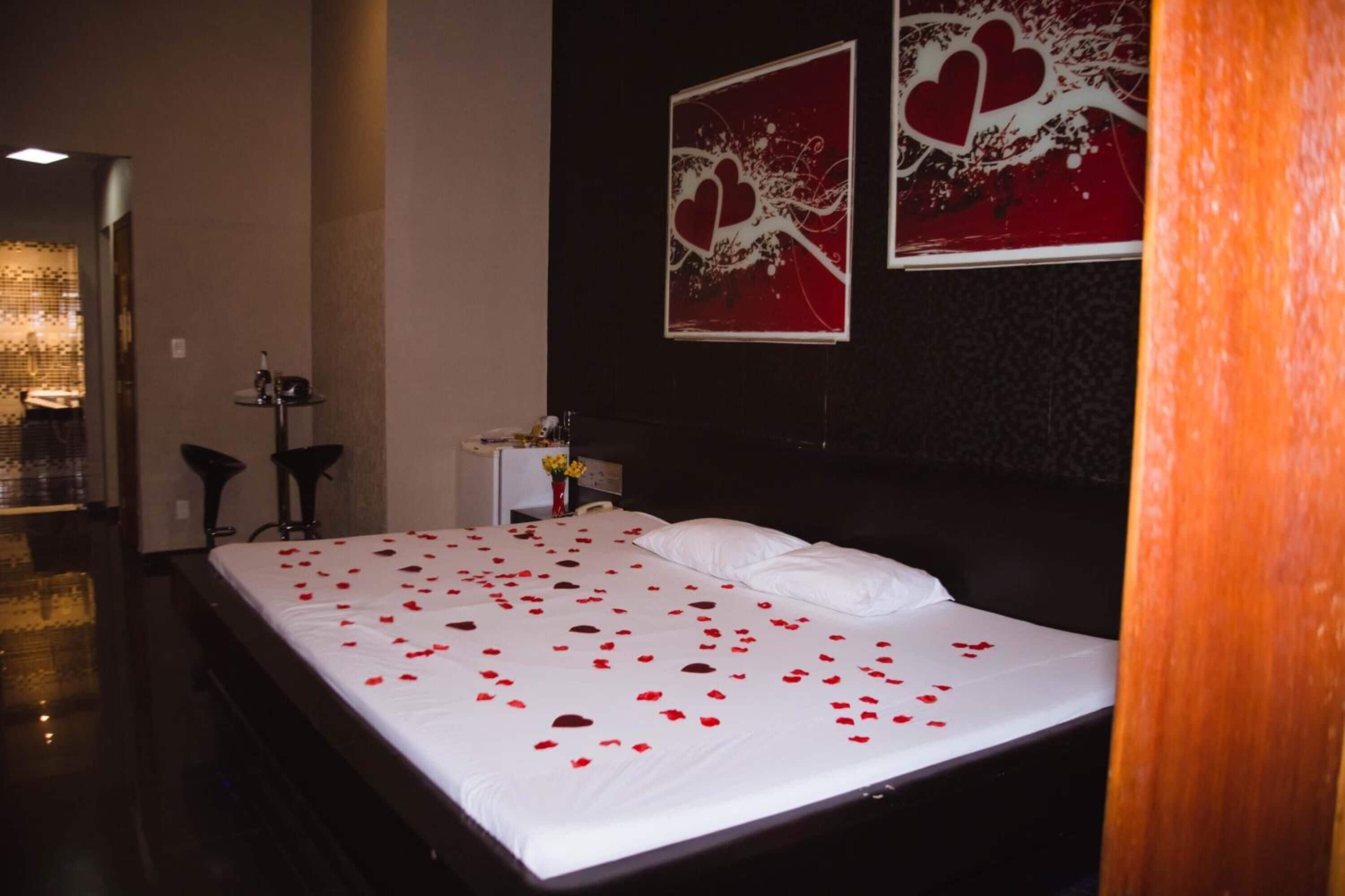 Una habitación de hotel con una cama cubierta de pétalos de rosa, una mesita de noche con un florero y dos cuadros abstractos con forma de corazón en la pared.