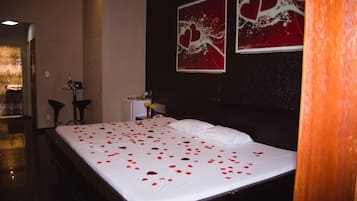 Una habitación de hotel con una cama cubierta de pétalos de rosa, una mesita de noche con un florero y dos cuadros abstractos con forma de corazón en la pared.