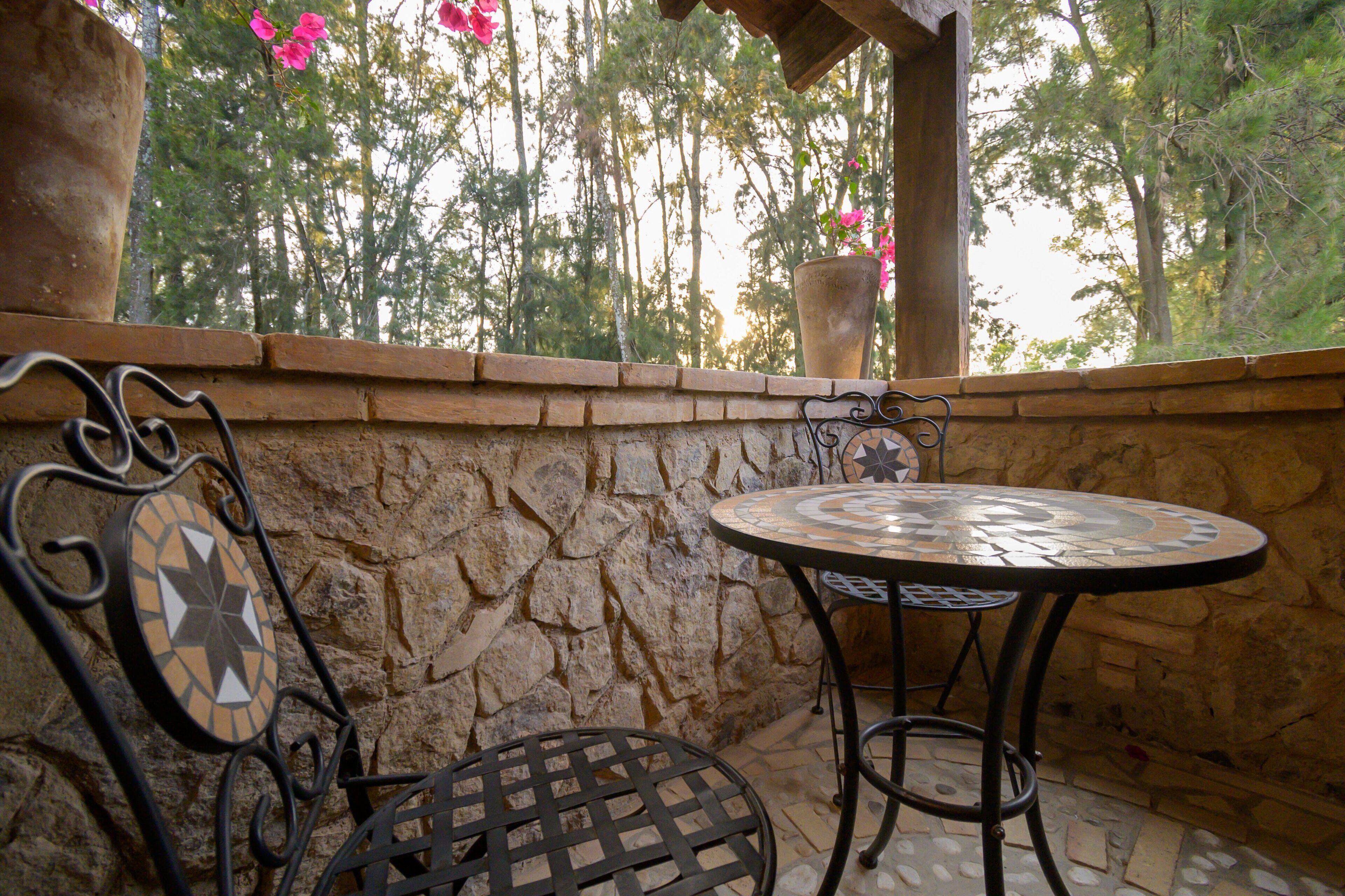 Un muro de piedra con una mesa y sillas con azulejos, una planta en maceta y vistas a árboles.