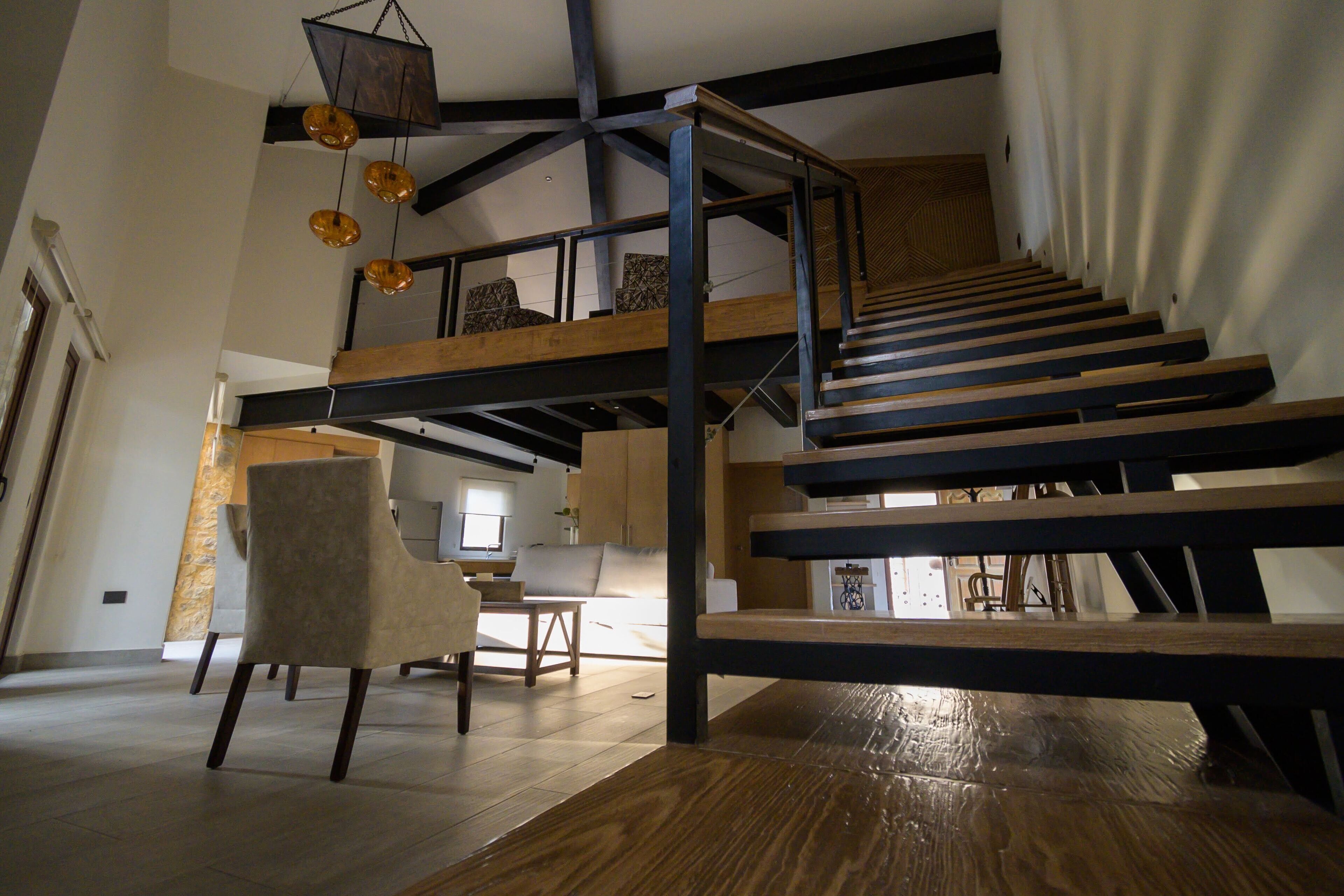 Un interior de varios niveles con vigas de madera a la vista, una escalera y un área de descanso con una silla y una mesa.