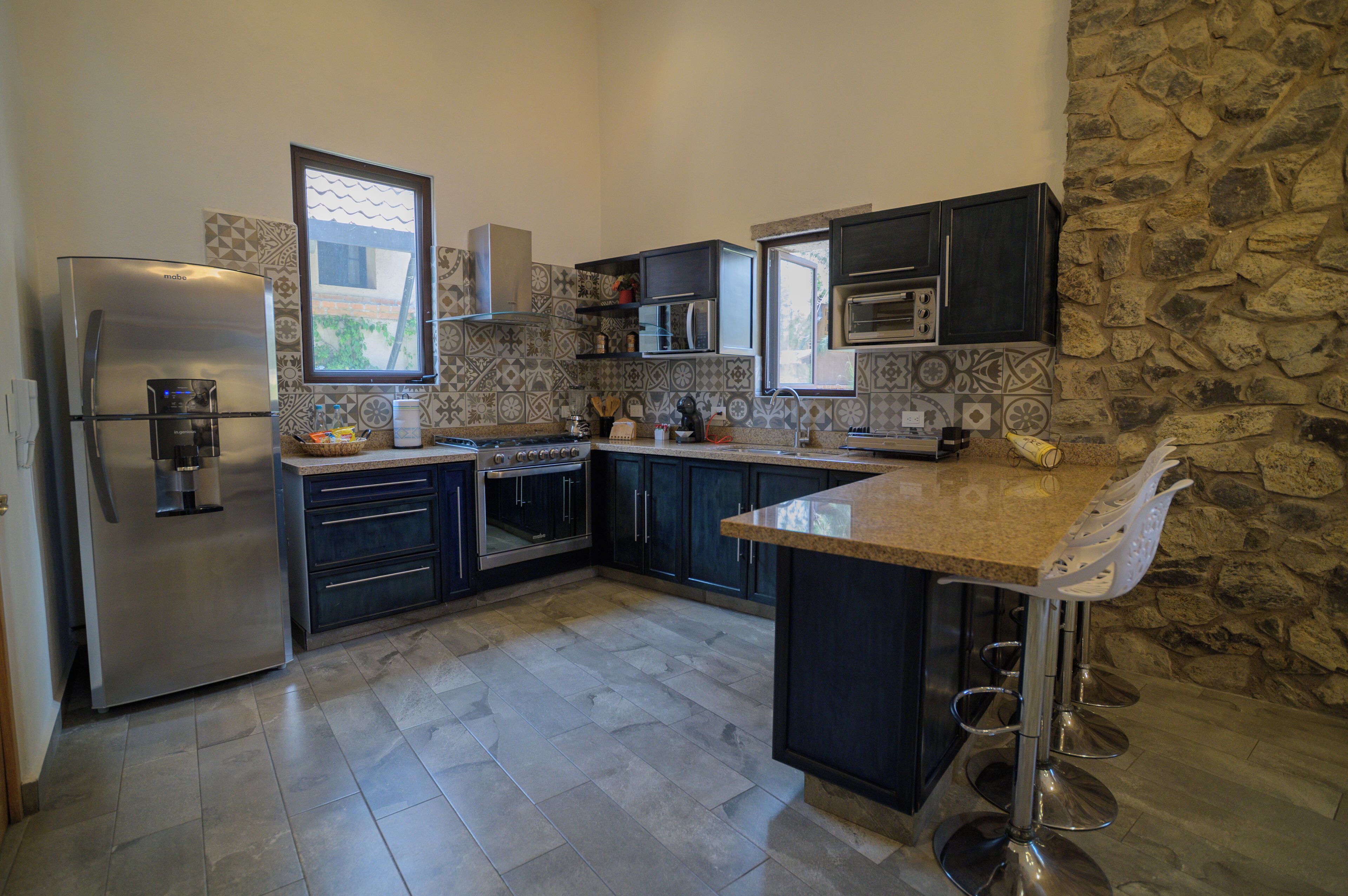 A modern kitchen with a built-in oven, microwave, and stainless steel refrigerator. It features dark cabinetry, a stone accent wall, and a large window providing natural light.