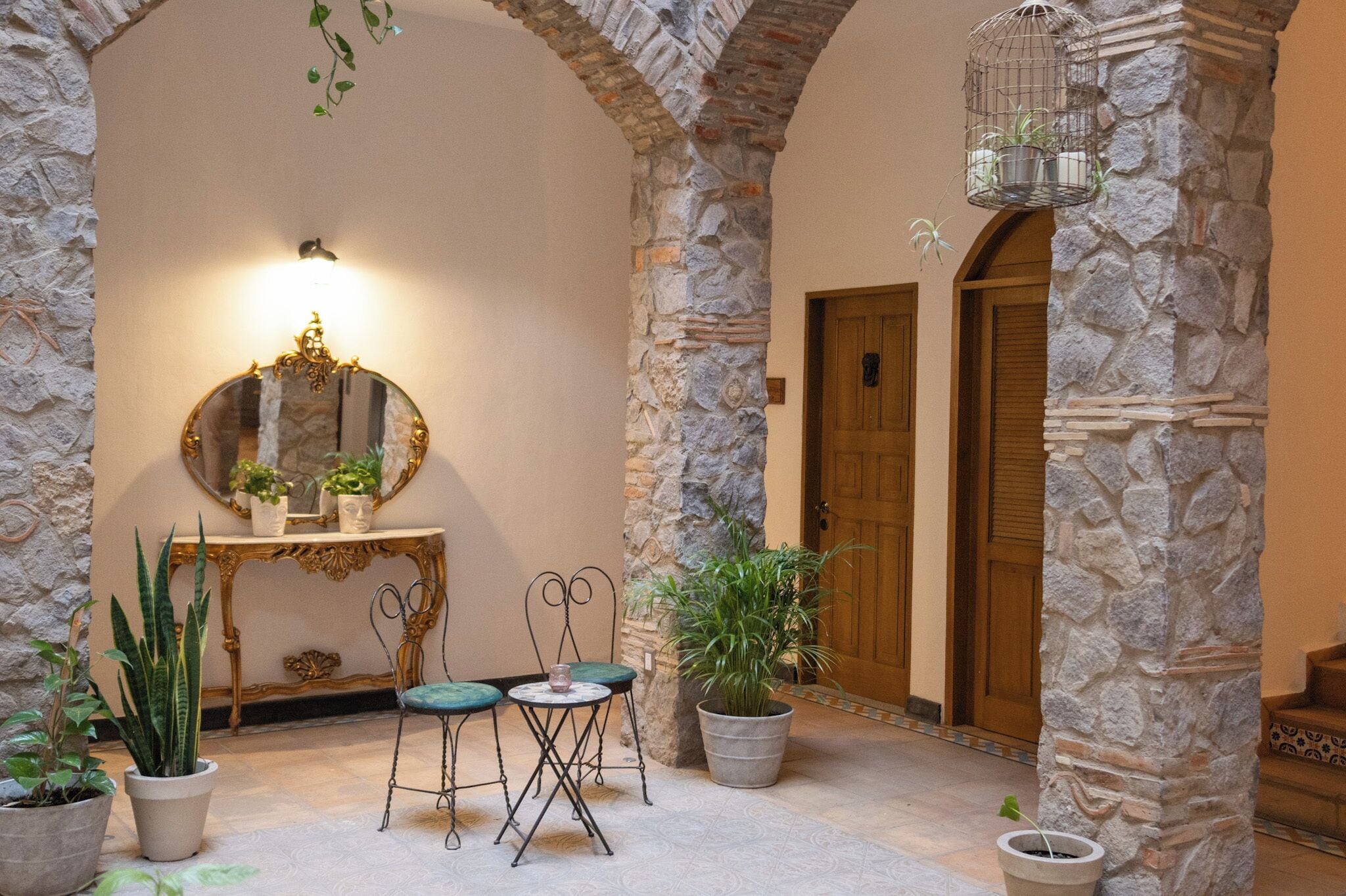 Un couloir aux murs de pierre, avec un miroir, une table entourée de chaises et des plantes en pot.