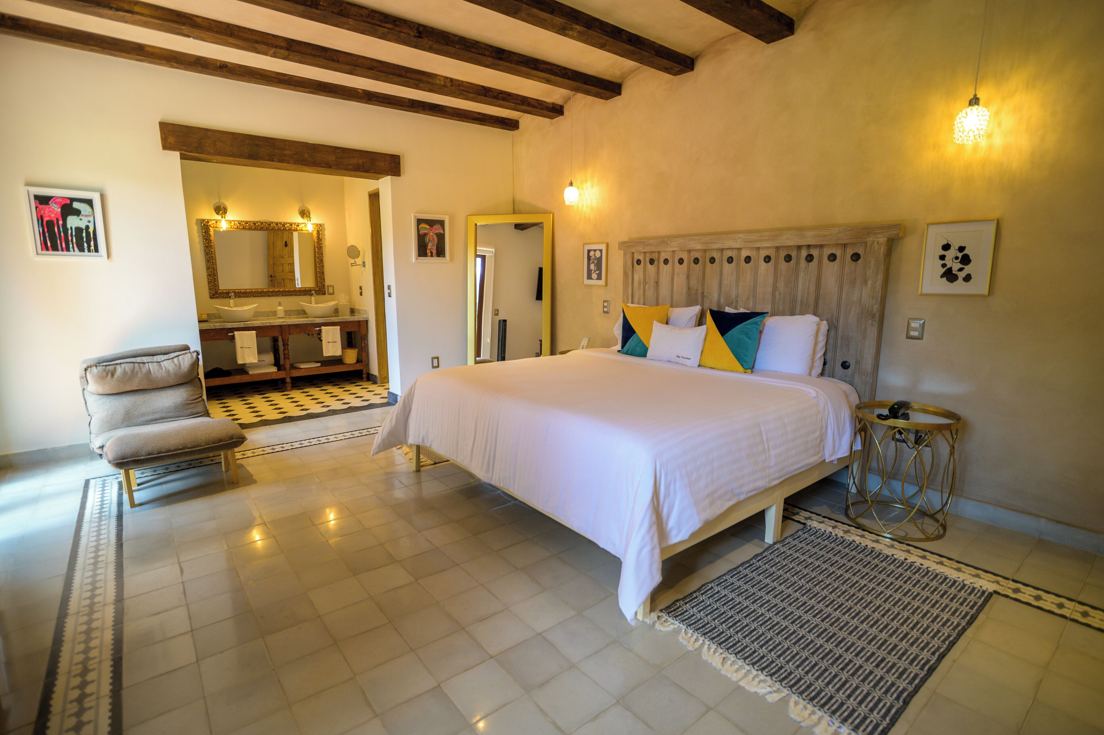A bedroom with a large bed, a sitting area with a chair, a vanity with a mirror, and a bathroom visible through an open door.