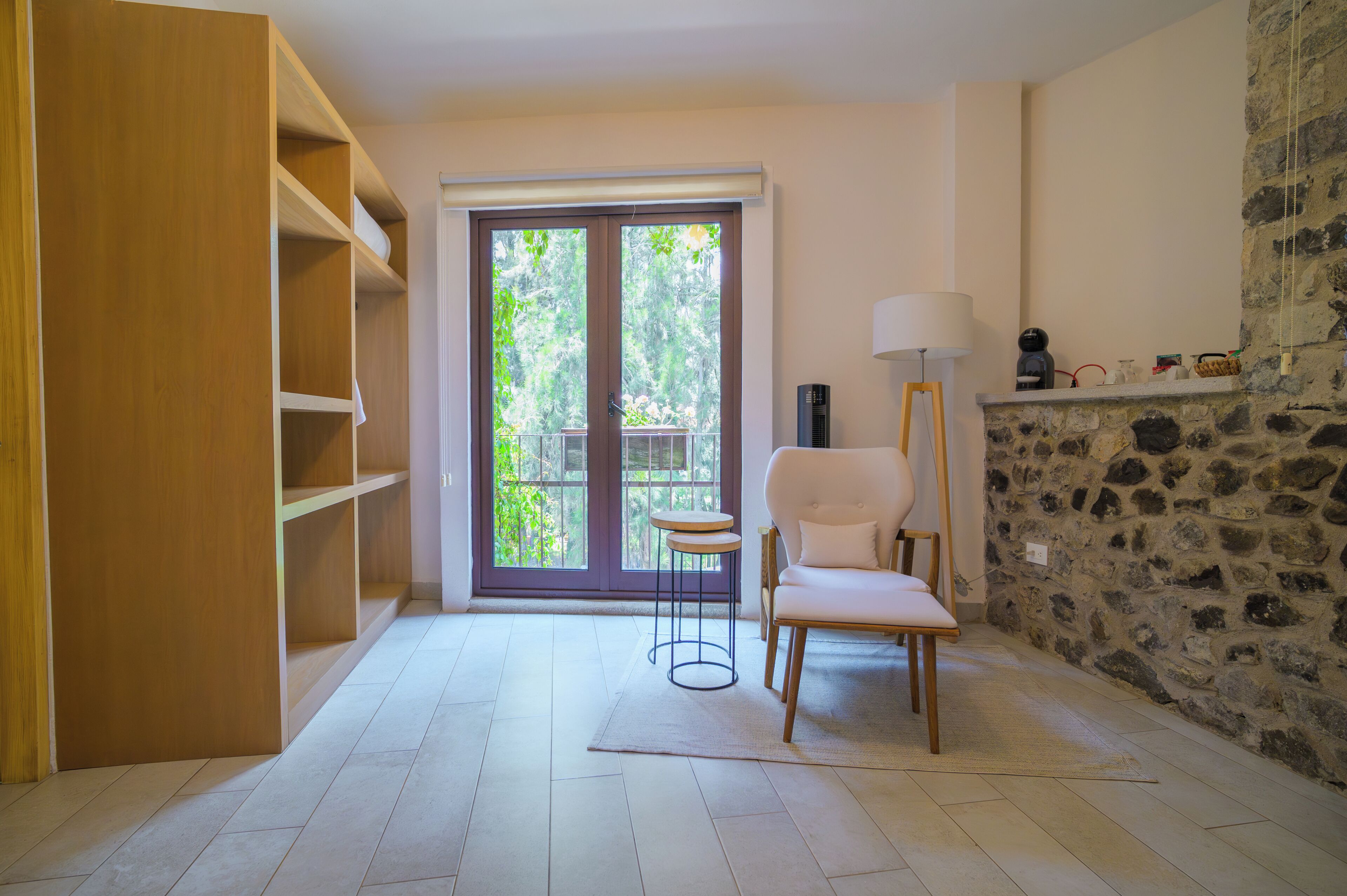 A room with a wooden wardrobe, a white chair, a small round table, a lamp, and a stone wall.
