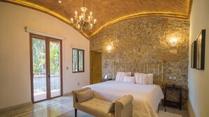 A bedroom with a large bed, a sofa, a chandelier, stone walls, and a view through a glass door.