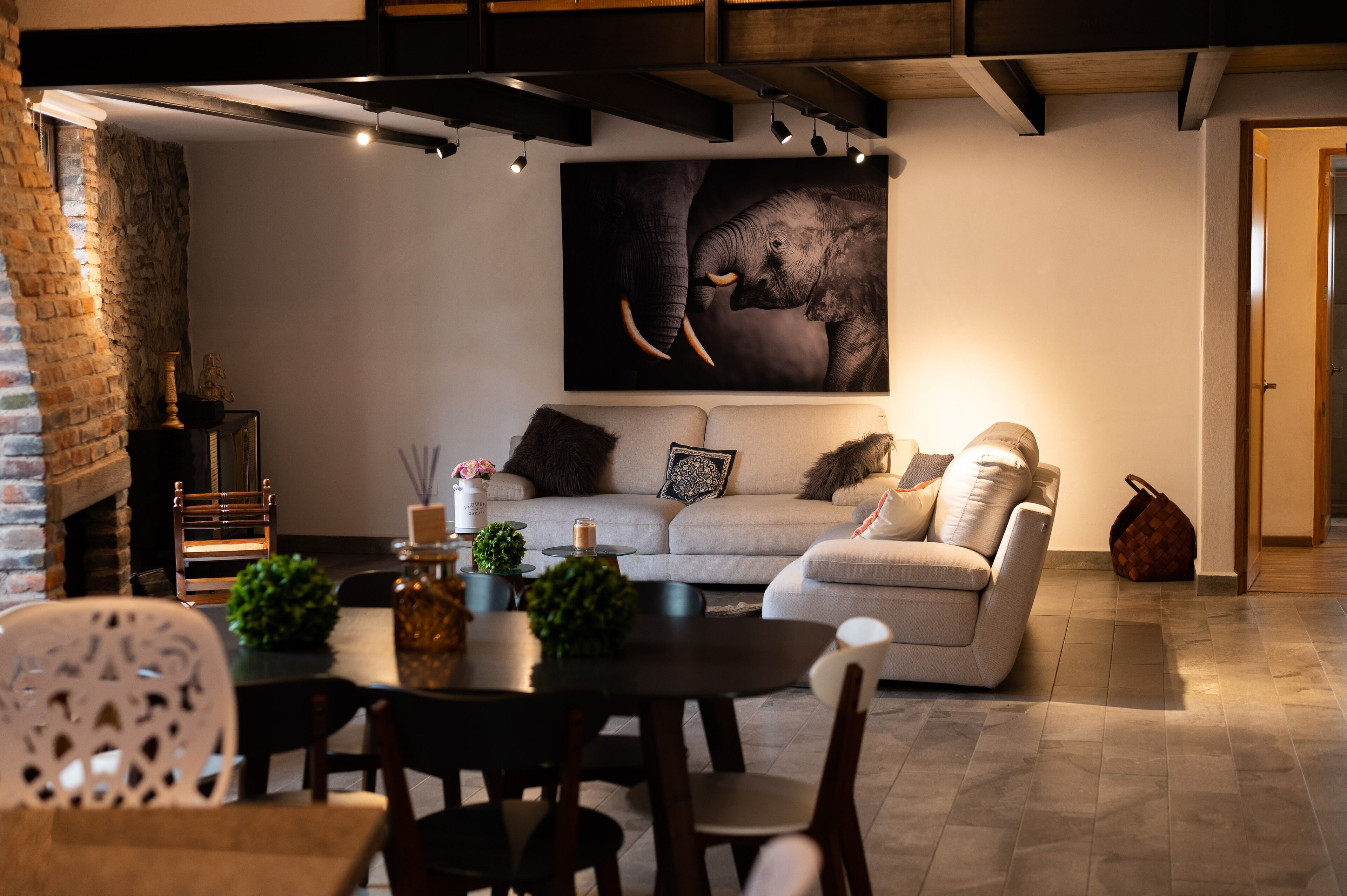 Un salon moderne avec un canapé, une table à manger et des chaises. On y trouve une cheminée, un grand tableau représentant un éléphant accroché au mur, et une porte qui donne sur une autre pièce.
